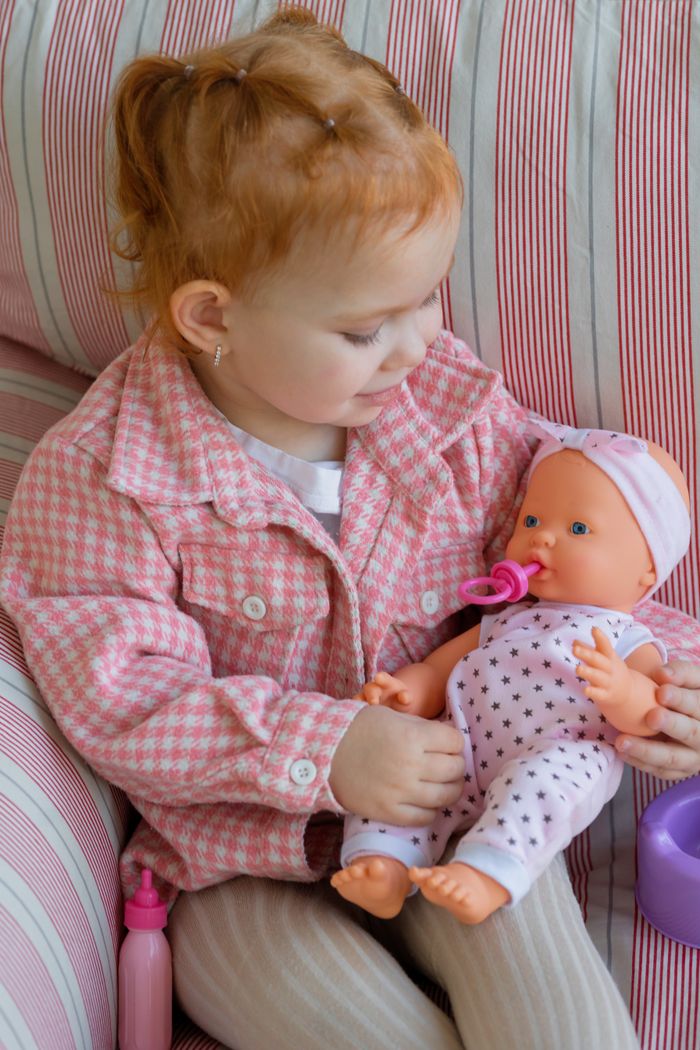 Little girl lovingly cradles a baby doll while seated on a stripe-patterned chair.